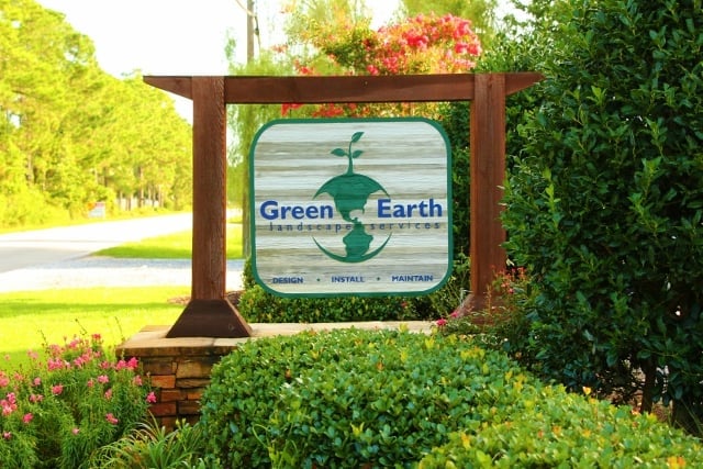 GreenEarth sign in on a stacked-stone base with shrubs, trees, and flowers surrounding it